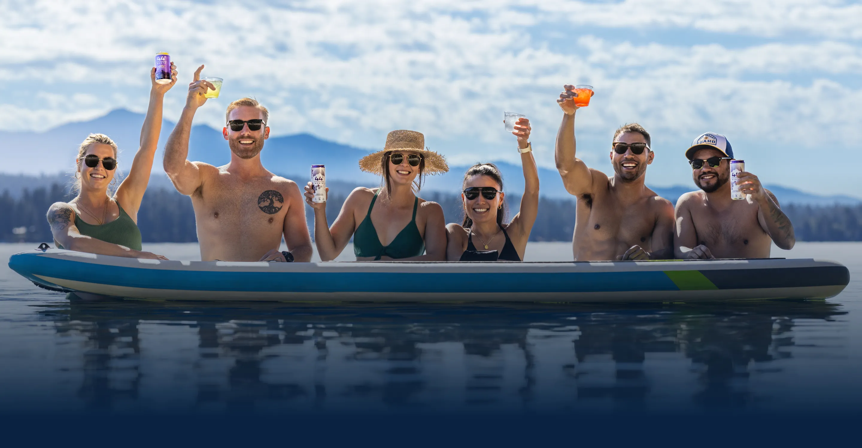Six friends raise drinks while sitting on a paddleboard on a calm lake with mountains in the background.